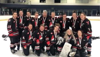 Photograph of a ladies, championship hockey team posing for a team photo at centre ice in a hockey rink, all players have medals around their necks 